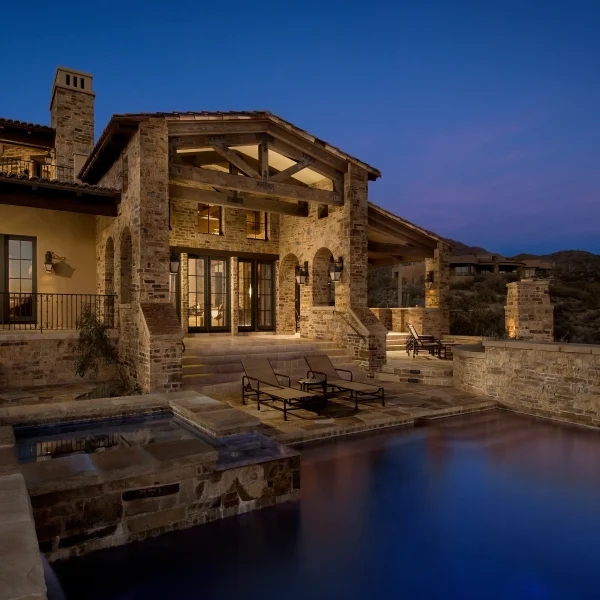 Stone villa with warm interior lights, timber beams, and a reflecting pool at dusk.