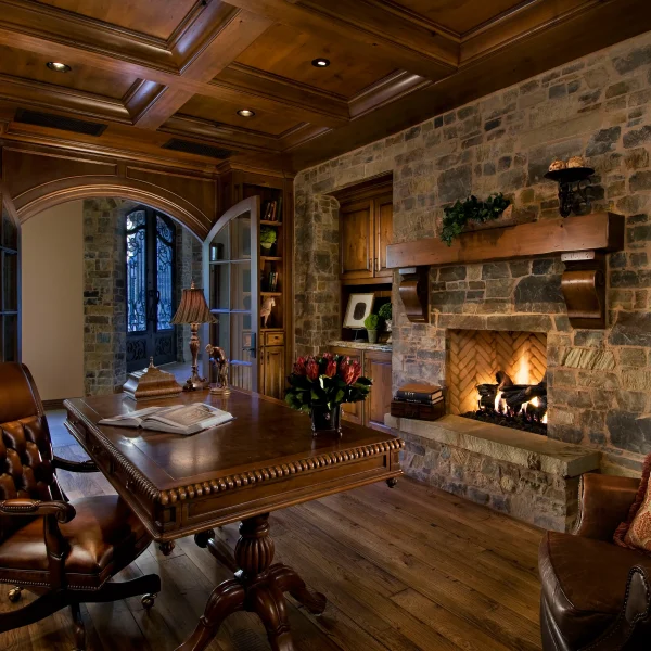 Cozy traditional study with a large wooden desk, leather chair, and built‑in bookshelves beside a stone fireplace.